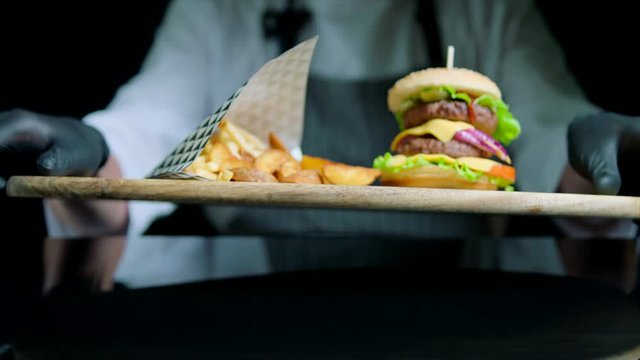Chef is serving set of tasty burger, french fries, beer on the wooden tray on the black background