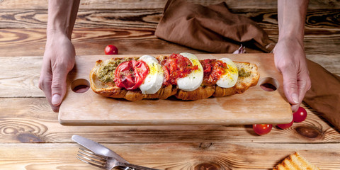 Female hands hold a cutting board with a bruschetta with baked tomatoes and mozzarella cheese.