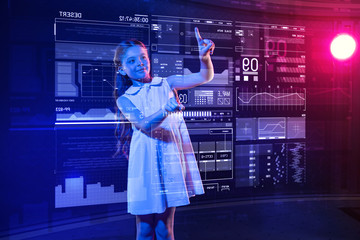 Pretty girl. Clever progressive schoolgirl doing her homework and smiling while using a futuristic transparent gadget