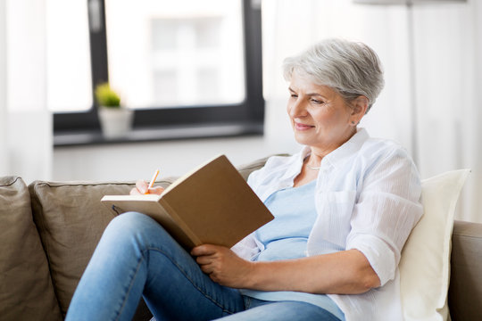 Age, Leisure And People Concept - Happy Senior Woman Writing To Notebook Or Diary At Home