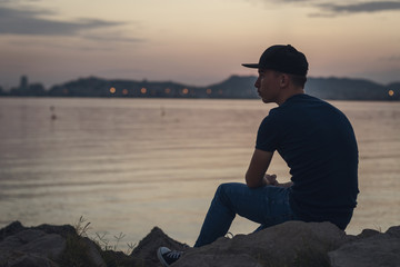 Pensive teen with black cap sitting on the rocks and looking to the sea