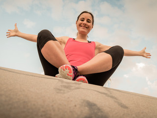happy fitness WOMAN after workout in pink shirt sitting on cement WITH arms up to a blue cloudy sky 