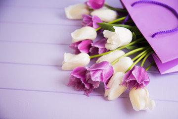 Beautiful bouquet of tender purple and white tulips in purple shopping bag on lilac wooden background.