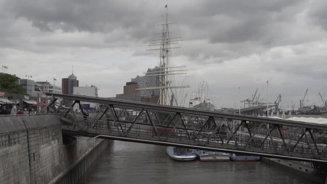 View at landing stage in Hamburg Harbour at day and person running down gangway with people walking on the promenade and masts of sailing boat Rickmer Rickmers and Elbphilharmonie in blurry backgound.