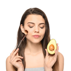 Beautiful young woman with avocado and cosmetic brush on white background