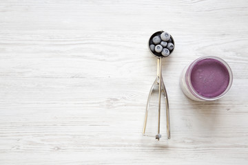 Blueberry ice cream sorbet in plastic jar, icecream scoop with frozen blueberries on a white wooden table, view from above. Top view, overhead. Copy space.