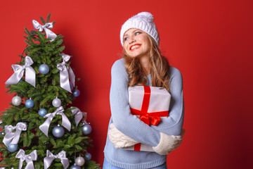 Beautiful young woman in hat and mittens with gift box near Christmas tree on color background