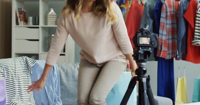 Beautiful caucasian young woman coming to the room, sitting in front of the camera, waving her heand and stsrting recording a gashion video blog. Inside.