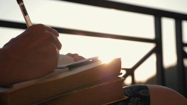 Woman's hand close-up, writing a note or letter to a notebook with a pen on a sunny day at sunset. A stack of scientific books, preparation for exams. Filling out the report