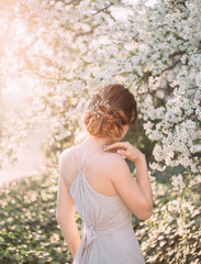 Red-haired girl in a modest, gray dress in rustic style. Portrait of the bride against the background of a flowering tree. A neat, collected hairdo in an airy beam. Photo from the back without a face