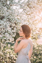 Red-haired girl in a modest, gray dress in rustic style. Portrait of the bride on the background of a blossoming tree with a minimal retouch. A neat, collected hairdo in an airy beam. Nude makeup.