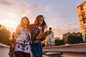 Smiling skate girls holding long-boards walking outdoors in the street having fun