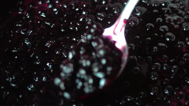 Hand stirs black currant jam in a saucepan with a metal spoon.