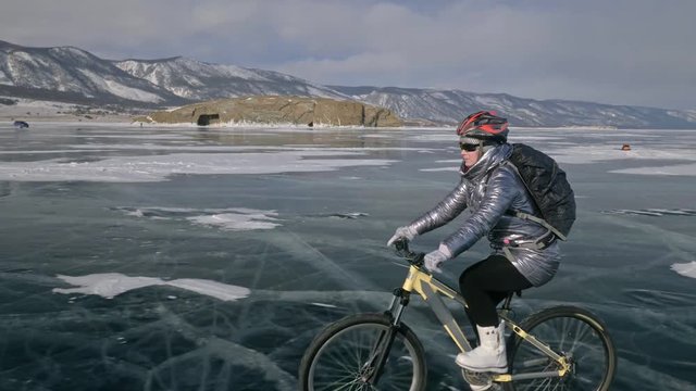 Woman is riding bicycle on the ice. The girl is dressed in a silvery down jacket, cycling backpack and helmet. Ice of the frozen Lake Baikal. The tires on the bicycle are covered with special spikes