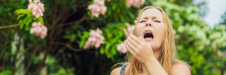 Young woman sneezes in the park against the background of a flowering tree. Allergy to pollen...
