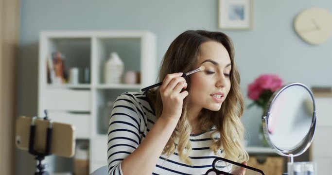 Young pretty woman shooting beauty blog video and doing makeup for eyes. Close up. Inside.