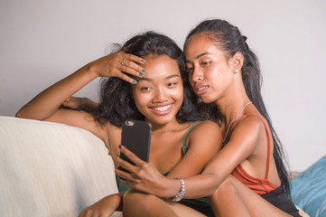 young happy and beautiful Asian sisters or girlfriends couple smiling cheerful taking selfie photo with mobile phone at home couch playful laughing together
