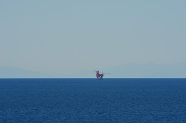 An oil rig, red in colour, is dwarfed by the vastness of the Mediterranean Sea. The sky is blue, with the faint outline of hills in the distance.