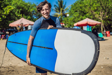Handsome sporty young surfer posing with his surfboard under his arm in his wetsuit on a sandy tropical beach