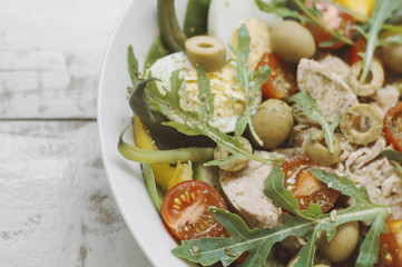 Nicoise salad with tuna fish and fresh vegetables. Bowl with canned tuna, eggs, arugula, spinach, bell pepper, cucumber, green beans, tomatoes and olives on wooden table