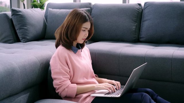 Young asian woman doing research work for her business. Smiling woman sitting on sofa relaxing while browsing online shopping website. Happy girl browsing through internet during free time at home.