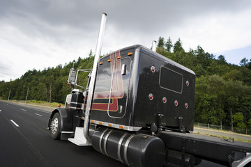Black classic big rig semi truck with vertical pipes going on wide multiline highway