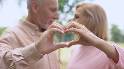 Positive aged couple showing heart sign by hands and hugging, tender relations