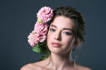 close-up portrait of attractive young woman with pink eustoma flowers behind ear looking at camera isolated on grey