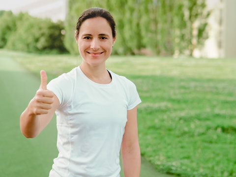happy smiling femal Fitness Coach in white sportswear making Thumbs up outdoor in green nature