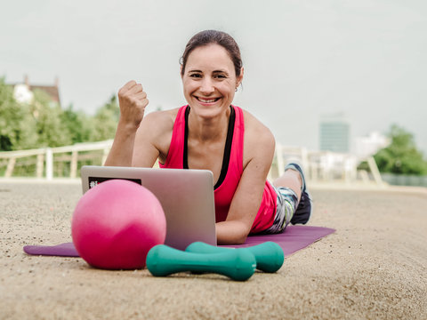 fit and sporty woman doing fitness workout online infront of a laptop on a yoga mat outside in the park 