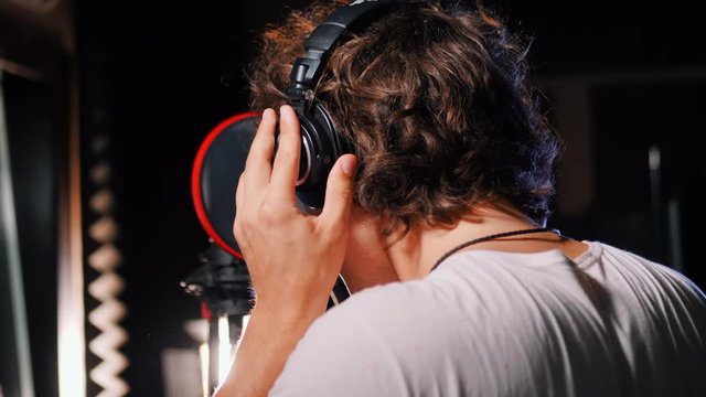 Young handsome singer man puts on headphones in the studio. Recording new melody or album. Male vocal artist with curly hair preparing for working. 4k