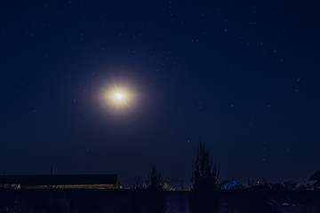 The moon and stars in the night sky over the roofs of village houses and trees.