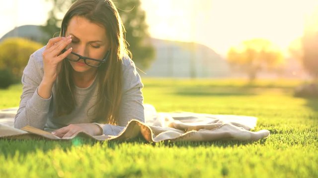 Girl in glasses reading book lying down on a blanket in the park at sunset