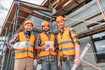 bottom view of group of smiling builders with blueprint and tablet looking at camera at...