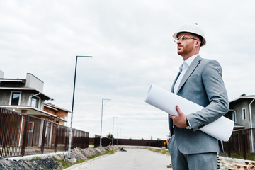 handsome architect in suit and hard hat holding blueprint looking at building house