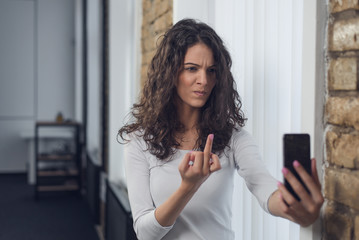 Angry young woman speaking on a mobile in office, making selfie, showing middle finger
