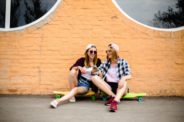 Two young happy girl friends in hipster outfit sitting on longboards and making selfie on phone