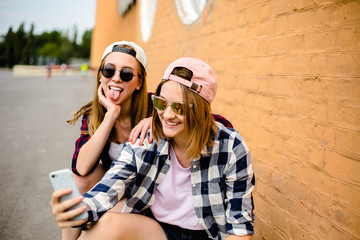 Two young happy girl friends in hipster outfit sitting on longboards and making selfie on phone