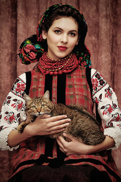 Naklejki Three quarter isolated portrait of a young Slavic woman in ethnic costume, wearing embroidered blouse, floral head scarf, red bead necklace, holding a cat on her lap, posing on dark brown background.