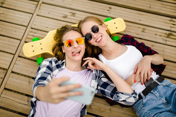 Two young girl in hipster outfit making selfie while lying with on wooden pier