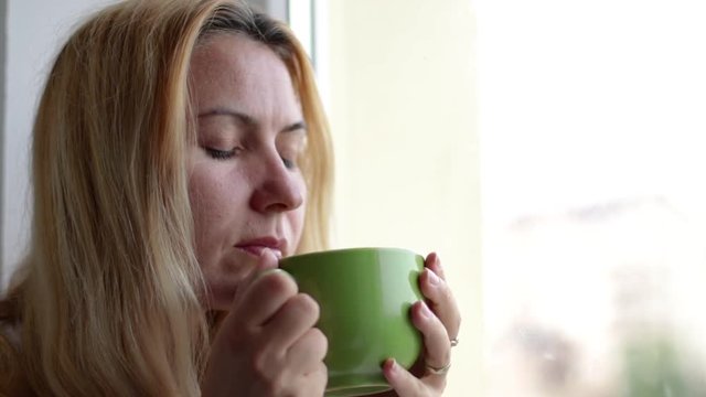Close up of woman sitting at the window and drinking a hot beverage. Blonde hair lady drinks hot tea
