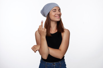 Happy woman with brown hair, wearing grey hat with cheerful expression, shows middle finger, has fun indoors, gestures at studio white wall. Attractive young female shows fuck sign. Body language.