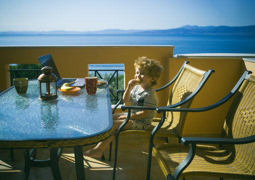 Sea holiday. Adorable kid sits at table in front of laptop and eats. Child having fast snack. Carbohydrates for children. Boy eating cookies. Family vacations at seaside. Sea or ocean at background.
