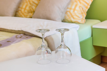 Two empty wine glasses turned upside down on night table next to the double bed in modern hotel bedroom. Valentines day or anniversary concept. Close up.