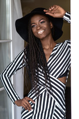 Attractive sexy girl with dark shiny skin posing for camera standing by the window touching her hat.