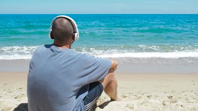 The man in the headphones is listening to music. A man on the beach listening to music.