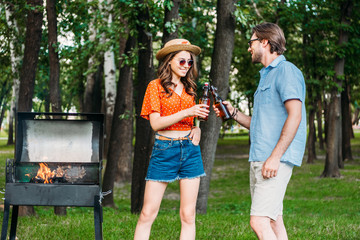young couple in sunglasses clinking bottles of beer during bbq in park