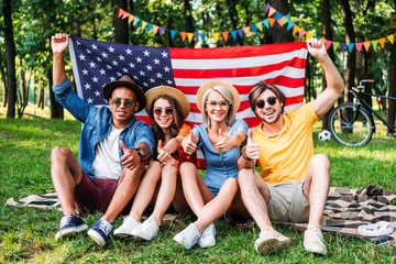 happy multiracial friends with american flag showing thumbs up in summer park