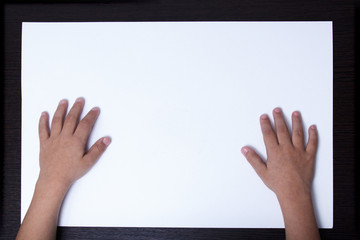 Hands of a child on a white sheet of paper on a wooden table.