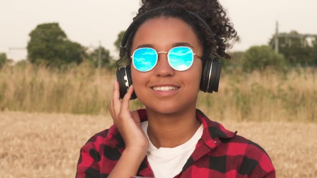 Slow motion video of beautiful mixed race African American girl teenager young woman wearing a red and black shirt and blue sunglasses listening to music on her cell phone and wireless headphones 